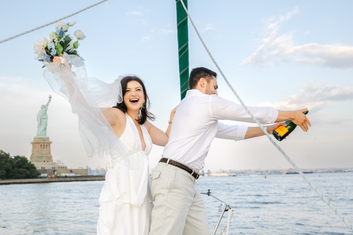 Couple celebrates on a sailboat near the Statue of Liberty, woman holds flowers, man opens champagne.