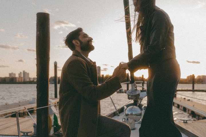 Man proposing to a woman on a boat at sunset, holding hands, with city skyline in the background.