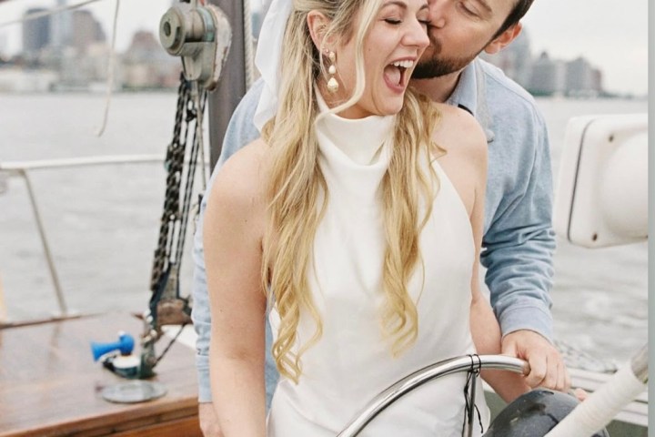 Couple on a sailboat, woman steering, man kissing her head, both smiling.