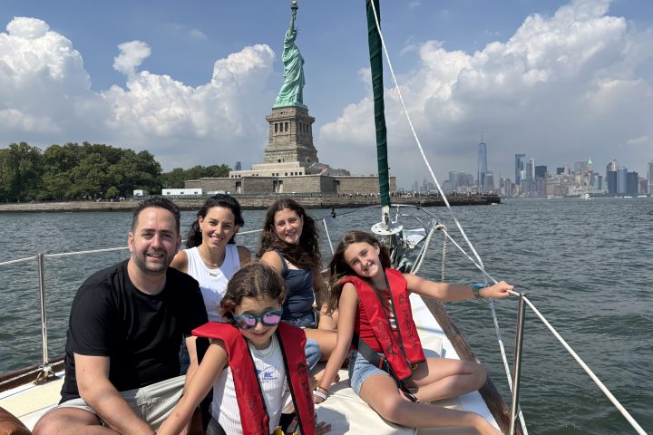 Group of people on a sailboat near the Statue of Liberty with a city skyline and cloudy sky in the background.