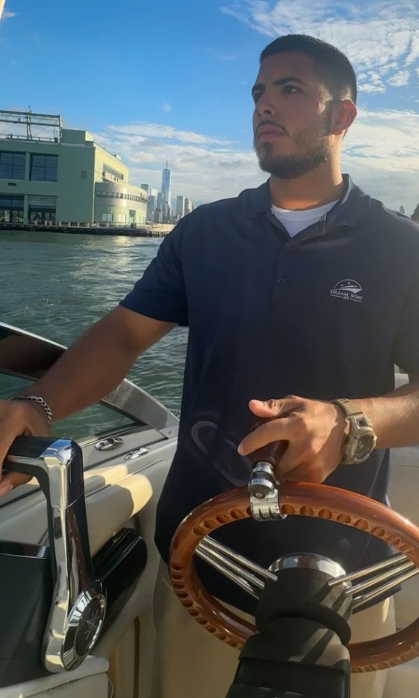 Man steering a boat on a sunny day with a city skyline in the background.