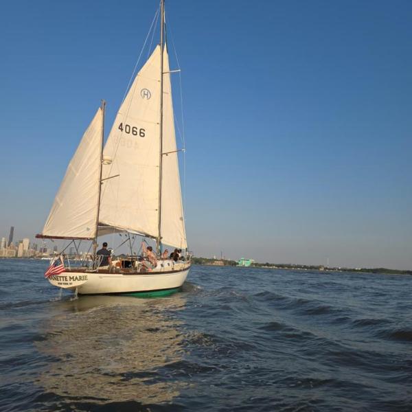 Sailboat with U.S. flag on calm water, city skyline in background.